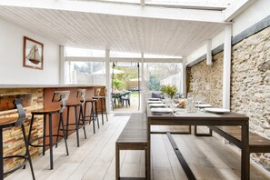 A modern dining area with wooden tables and bar stools, a stone wall, and a view of the outdoors. - House with garden for 8, 550m from the beach (Barbâtre)