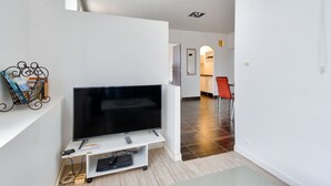 Un saló modern amb un televisor de pantalla plana sobre un suport blanc, una petita estant amb llibres i un objecte decoratiu, i una cuina al fons.