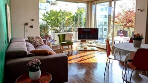 Un salon moderne comprenant un canapé, une table basse et un téléviseur, de grandes fenêtres offrant une vue sur les arbres et les bâtiments, ainsi qu’un coin repas avec des chaises et une table.