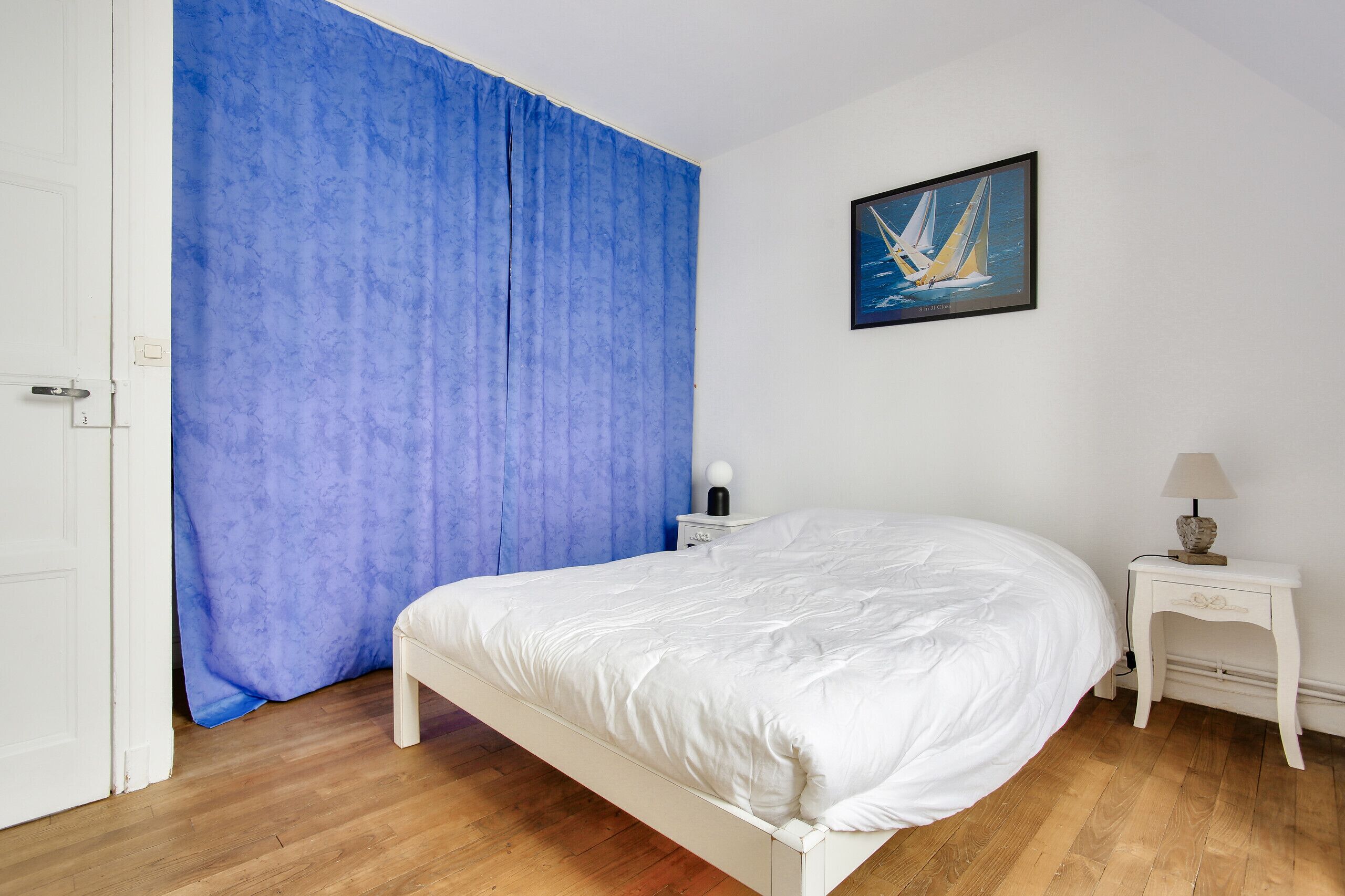 Un dormitorio con cama blanca, cortina azul, mesita de noche con lámpara y un cuadro de un velero en la pared.