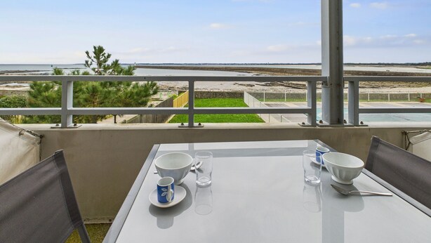 Un balcón con una mesa dispuesta para dos, con vista a una playa y una costa distante.