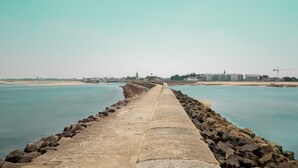 Un largo y recto muelle de piedra que se adentra en una masa de agua tranquila, con cielo despejado y edificios a lo lejos.