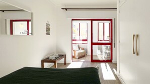 Une chambre à coucher comprenant un lit, une petite table, un banc et une porte-fenêtre coulissante donnant sur un balcon.