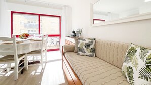 Une pièce lumineuse comprenant une table à manger, des chaises, un canapé et une grande fenêtre aux montants rouges.