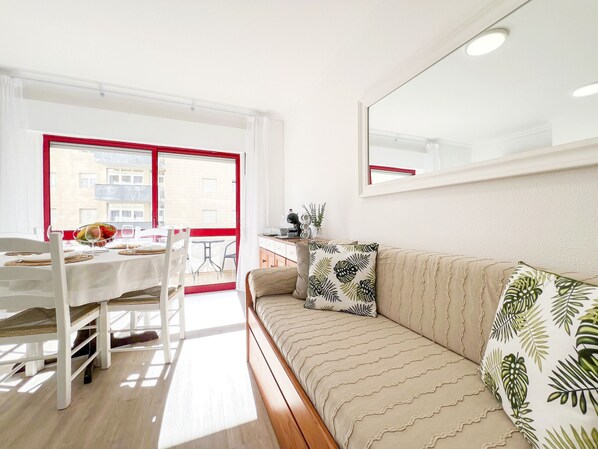 A bright room with a dining table, chairs, a sofa, and a large window with red frames.