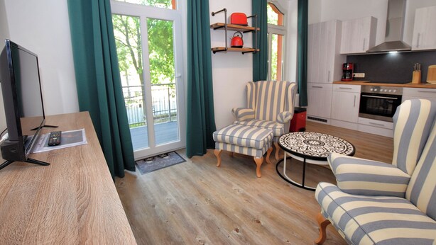 Ein Wohnzimmer mit Fernseher, gestreiften Sesseln, einem kleinen Tisch und einer Küchenzeile mit Schränken und Geräten.