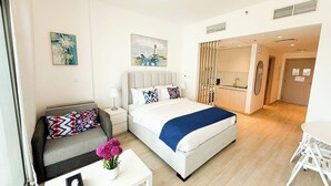 A modern hotel room with a bed, a sofa, a small table with a vase of flowers, a painting on the wall, and a kitchenette in the background. - Yas Island Waters Edge Studio (Abu Dhabi)