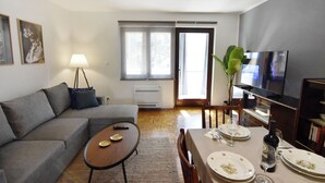 A living room with a sofa, a coffee table, a dining table set for two, a TV, and a large window with curtains.
