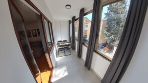 A narrow corridor with a dining area featuring a table and chairs, large windows with curtains, and a view of a tree and buildings outside.
