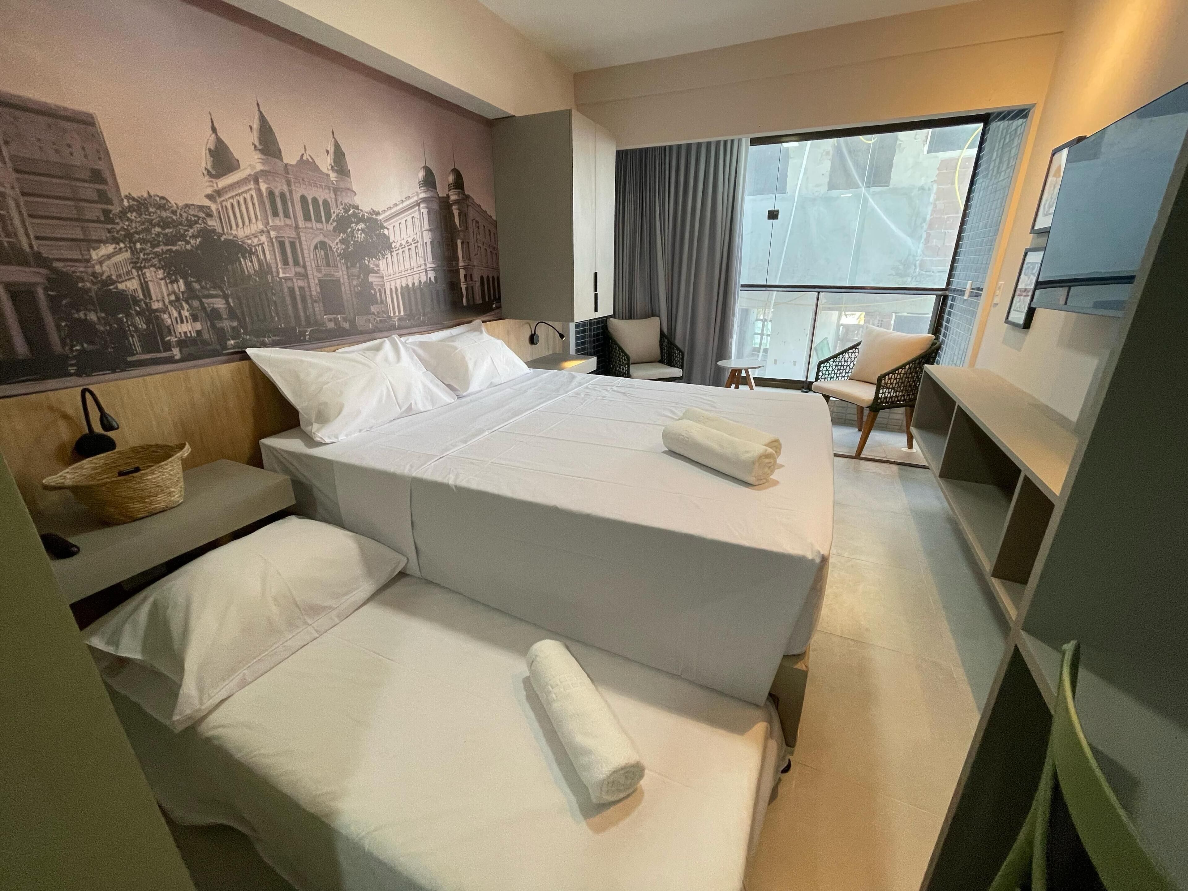 A hotel room with a large bed, a small desk, a chair, and a view of a cityscape through a large window.
