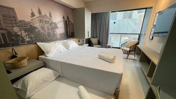 A hotel room with a large bed, a small desk, a chair, and a view of a cityscape through a large window.