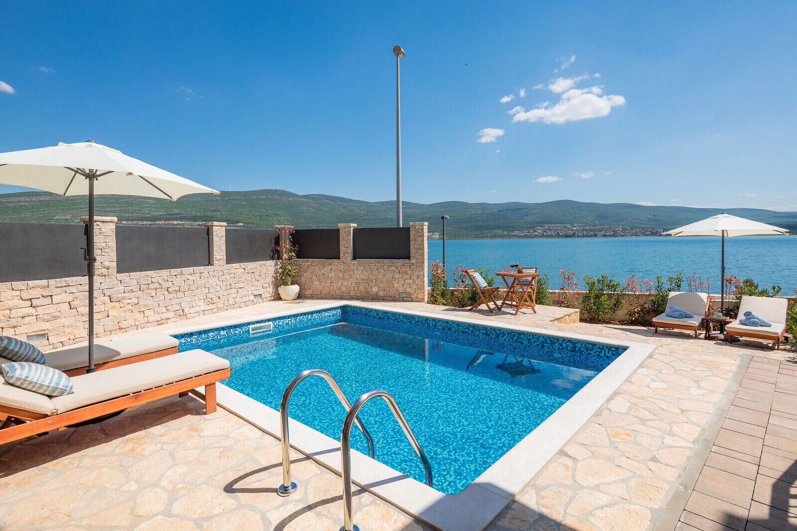 Área de piscina com vista para o mar, paredes de pedra e espreguiçadeiras.