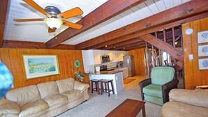A cozy living room with a sofa, a wooden bench, and a fan. A kitchen is visible in the background with a microwave, stove, and refrigerator. - Village West 1-Baywater 28th St, Free WiFi, W/D, AC (Ocean City)