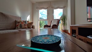 Een woonkamer met een houten vloer, een gestreept voetenbankje, een bank met bloemenkussens, een fauteuil, een televisie en een raam met uitzicht op groen.