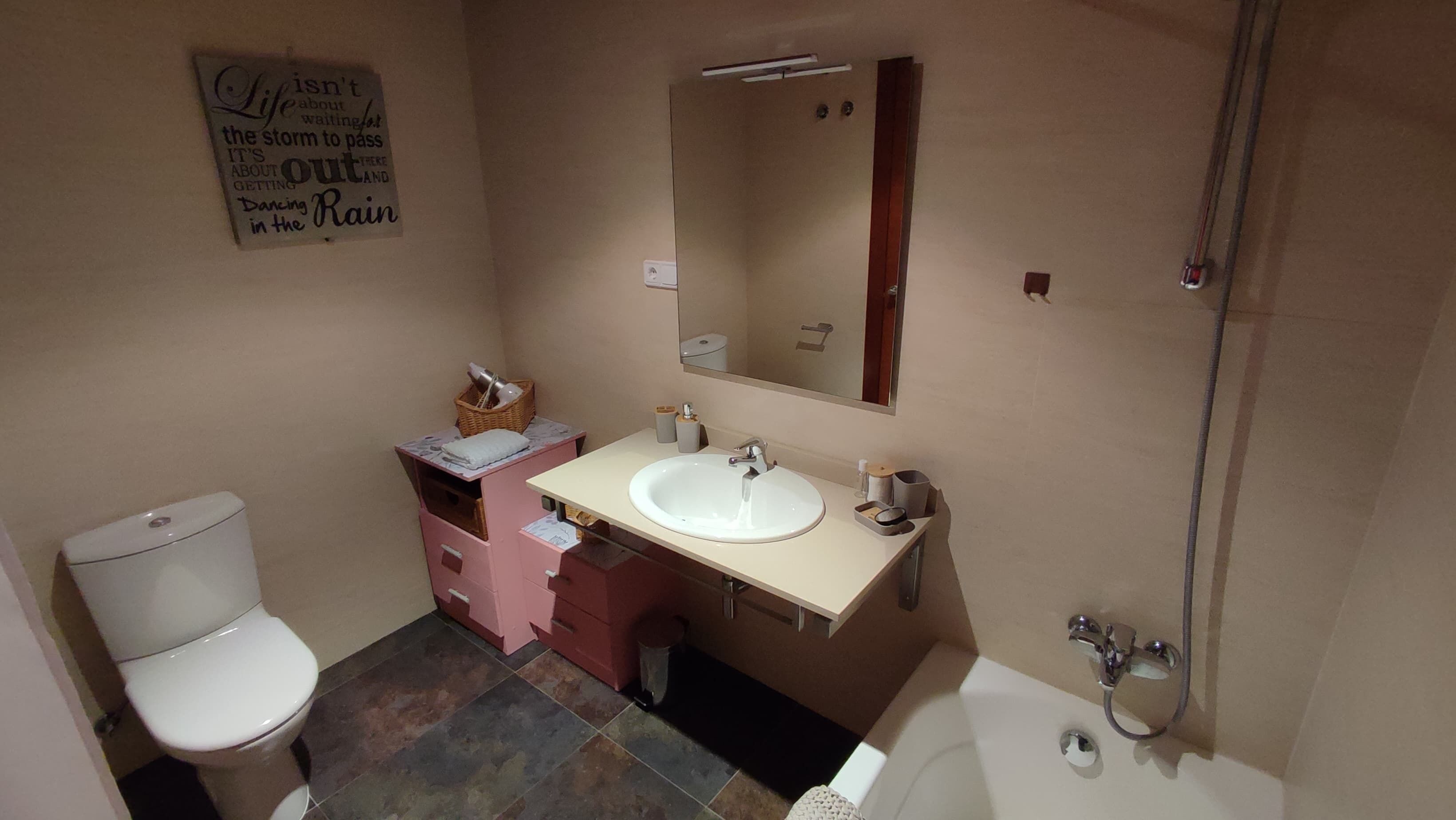 Un baño con inodoro, lavabo y bañera. Hay un espejo sobre el lavabo y un perchero de toallas en la pared. Un cartel en la pared dice: 'Life isn't about the storm to pass, it's about getting out and dancing in the rain'.