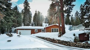 A wooden cabin with a garage, surrounded by snow-covered ground and tall evergreen trees. - Tahoe Trail Retreat Steps From the TRT (Stateline)