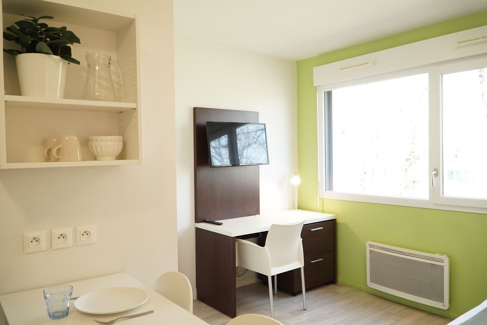 Una habitación moderna con escritorio, silla y televisor montado en la pared. Hay una ventana que proporciona luz natural y un radiador debajo.