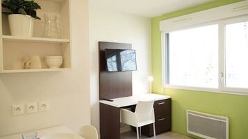 Una habitación moderna con escritorio, silla y televisor montado en la pared. Hay una ventana que proporciona luz natural y un radiador debajo.