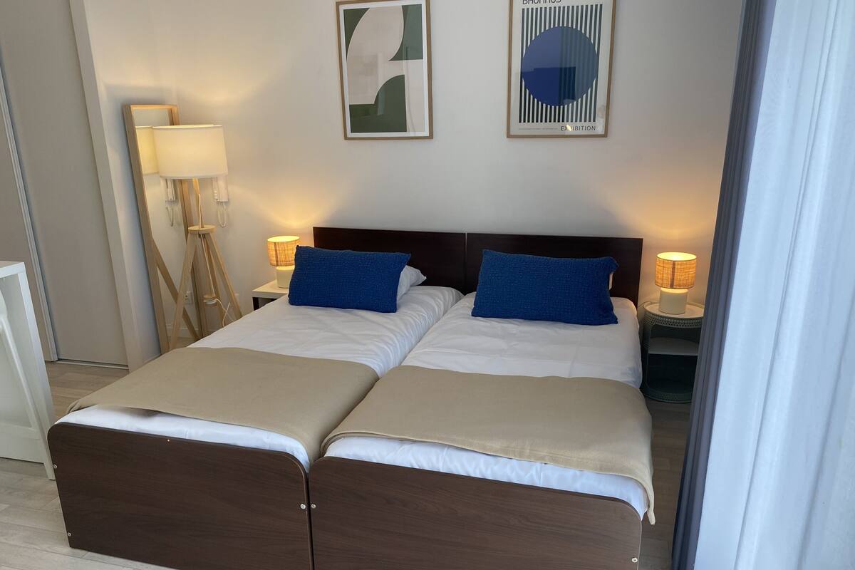 Cama doble con estructura de madera, dos almohadas azules y una manta color beige. Hay dos mesitas de noche con lámparas, y una obra de arte en la pared.