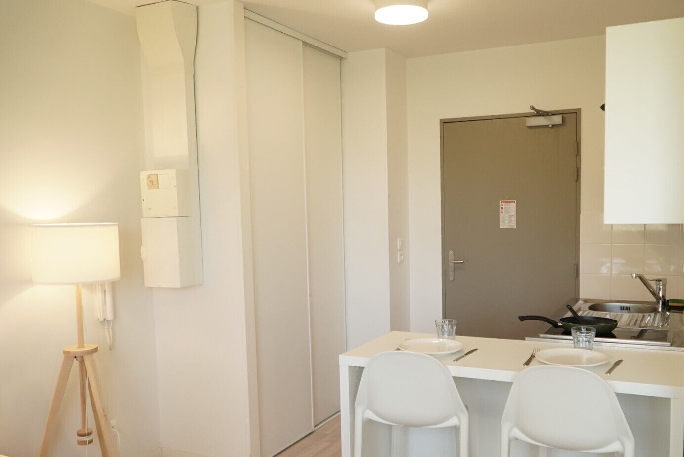 A small, modern kitchen with a dining table and two chairs, a single light source, and a door with a sign.