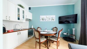 A modern kitchen with white cabinets, a round dining table with chairs, a flat-screen TV mounted on the wall, and a small side table with a fan and books. - Luxury European Apartment (Kraków)