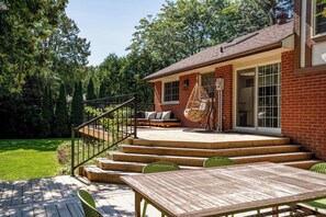 A wooden deck with a table and chairs, a hanging chair, and a staircase leading to a covered porch with a brick wall and large windows.