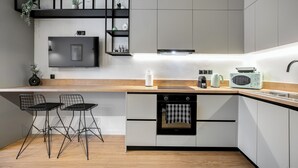 Eine moderne Küche mit Einbauofen, Mikrowelle und Kaffeemaschine. Zwei schwarze Stuhlgestelle aus Draht und eine Holz Arbeitsplatte sind vorhanden.