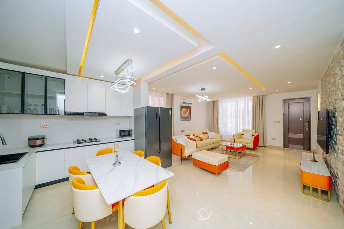 A modern apartment with a kitchen, dining area, and living room. It features a marble table, yellow chairs, a sofa set, and a TV mounted on the wall.