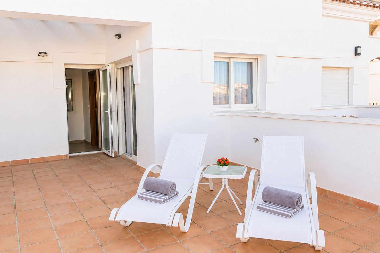 Een balkon met twee witte loungesteilen, een kleine tafel met een bloevas, en een schuifdeur die leidt naar een binnenruimte.
