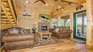 A cozy cabin interior with a fireplace, two leather sofas, a coffee table, and a wooden staircase. - Wild Honey- 4bdrm, Incredible Mtn Views, Pool Table, Fire Pit, Pool Access (Sevierville)