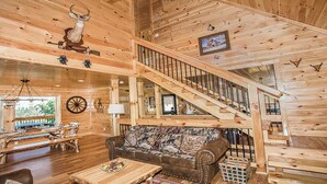A wooden cabin interior with a staircase, a couch with animal prints, a dining area, and a mounted deer head. - Wild Honey- 4bdrm, Incredible Mtn Views, Pool Table, Fire Pit, Pool Access (Sevierville)
