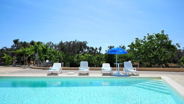 A swimming pool with clear water, surrounded by white lounge chairs and a blue umbrella.