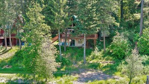 A cabin with a deck and a small porch, surrounded by dense forest and a body of water. - Rustic A-Frame Cabin | Dock | Paddleboards | Summer Camp Vibes (Coeur D'Alene)