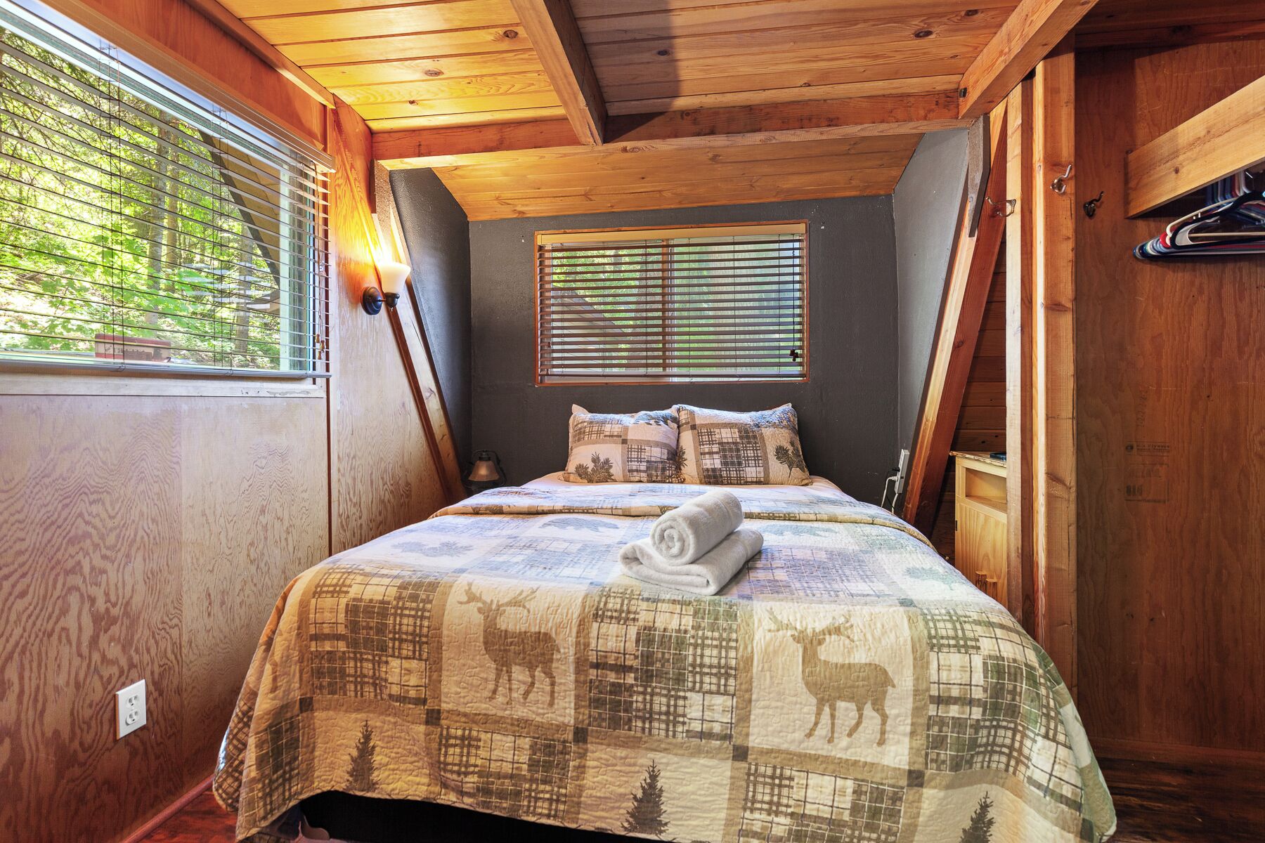 A cozy bedroom with a wooden ceiling, a bed with a quilt featuring animal prints, two bedside lamps, and a window with blinds.