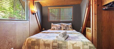 A cozy bedroom with a wooden ceiling, a bed with a quilt featuring animal prints, two bedside lamps, and a window with blinds.