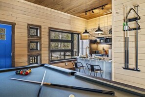 Une table de billard dans une pièce aux murs en bois, avec un coin cuisine visible à travers une fenêtre.