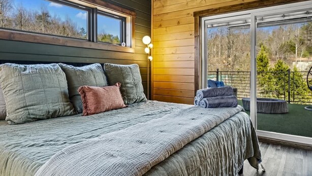 Een slaapkamer met een groot bed, meerdere kussens, een nachtkastje en een raam met uitzicht op bomen.