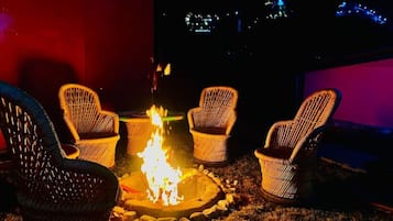 Un espacio al aire libre con mesas y sillas de mimbre dispuestas alrededor de un brasero central.