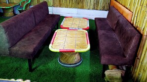 A room with two sofas and two square tables with woven tops, a bamboo wall, and artificial green carpet. - The Escapee Resort (Dhanaulti)