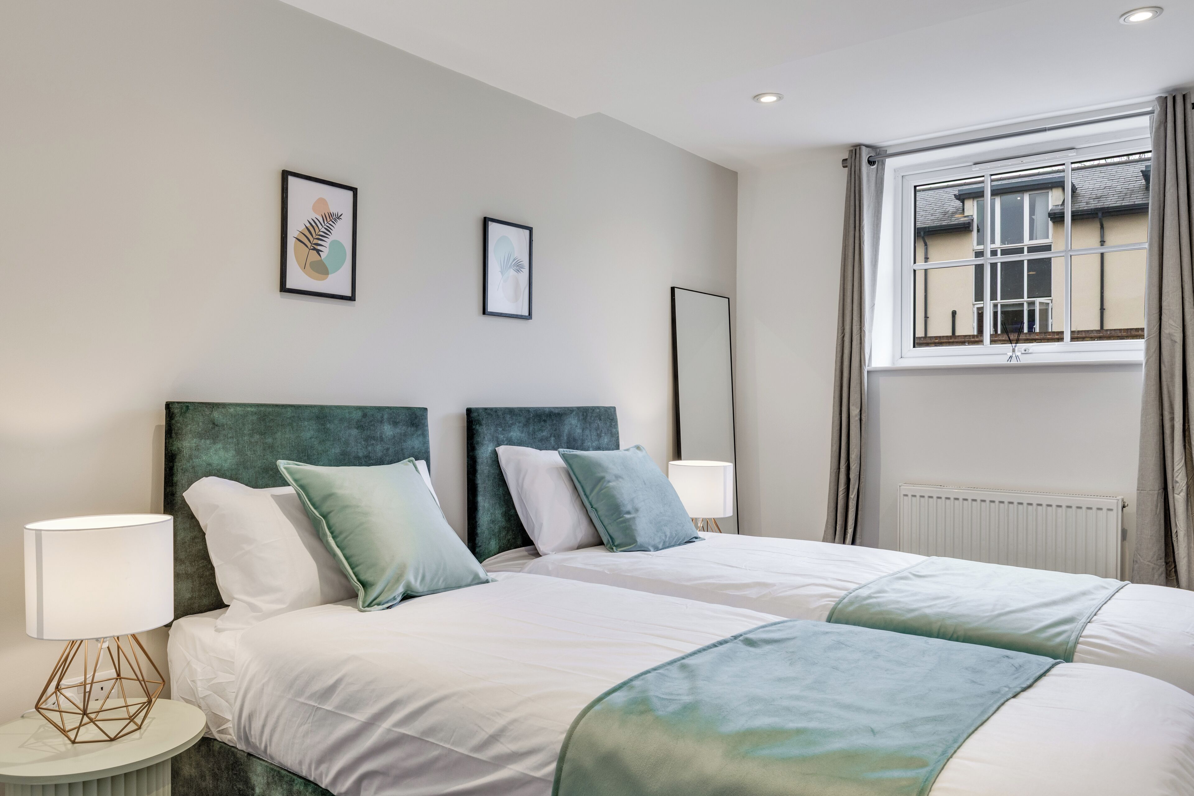 A double bed with a green headboard, white bedding, and two bedside lamps. There are two framed artworks on the wall and a window with curtains.
