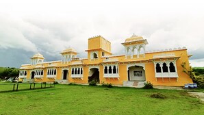 Un bâtiment jaune de style traditionnel, doté de plusieurs dômes et de fenêtres en arc de cercle, entouré d’une zone verdoyante.