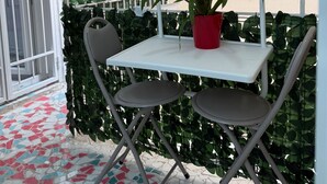 Un pequeño patio al aire libre con una mesa blanca y dos sillas, una planta en maceta roja y una pared verde cubierta de hojas.