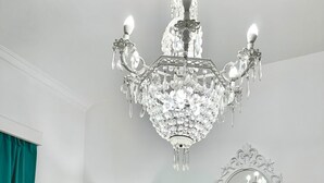 Un dormitorio con una cama blanca ornamentada, almohadas verdes, una mesita de noche blanca, una lámpara verde, un espejo blanco y una araña de cristal.
