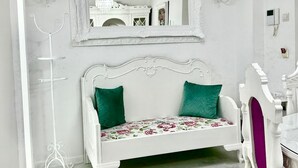 Un sofá blanco con cojines verdes y tapicería floral, un perchero metálico blanco y un espejo blanco ornamentado en la pared.