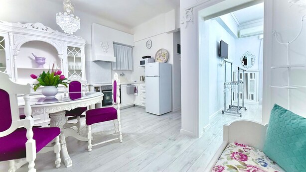 Una cocina moderna con armarios blancos, un comedor con una mesa blanca y sillas moradas, y una sala de estar con un sofá color turquesa.