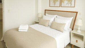 A neatly made bed with a beige bedspread and pillows, a bedside table with a lamp, and two framed floral artworks on the wall. - Residence del Maestro Apartments (Asunción)