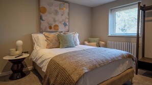 Una camera da letto con un letto, cuscini, un comodino, un quadro appeso al muro, una finestra con persiane e un termosifone.