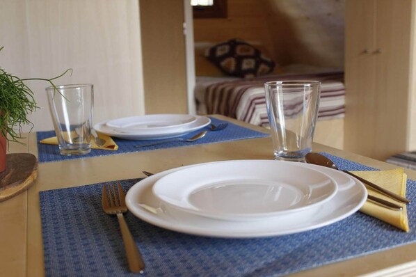 Una mesa de comedor dispuesta con platos, cubiertos y vasos, con una planta y una cama a lo lejos.