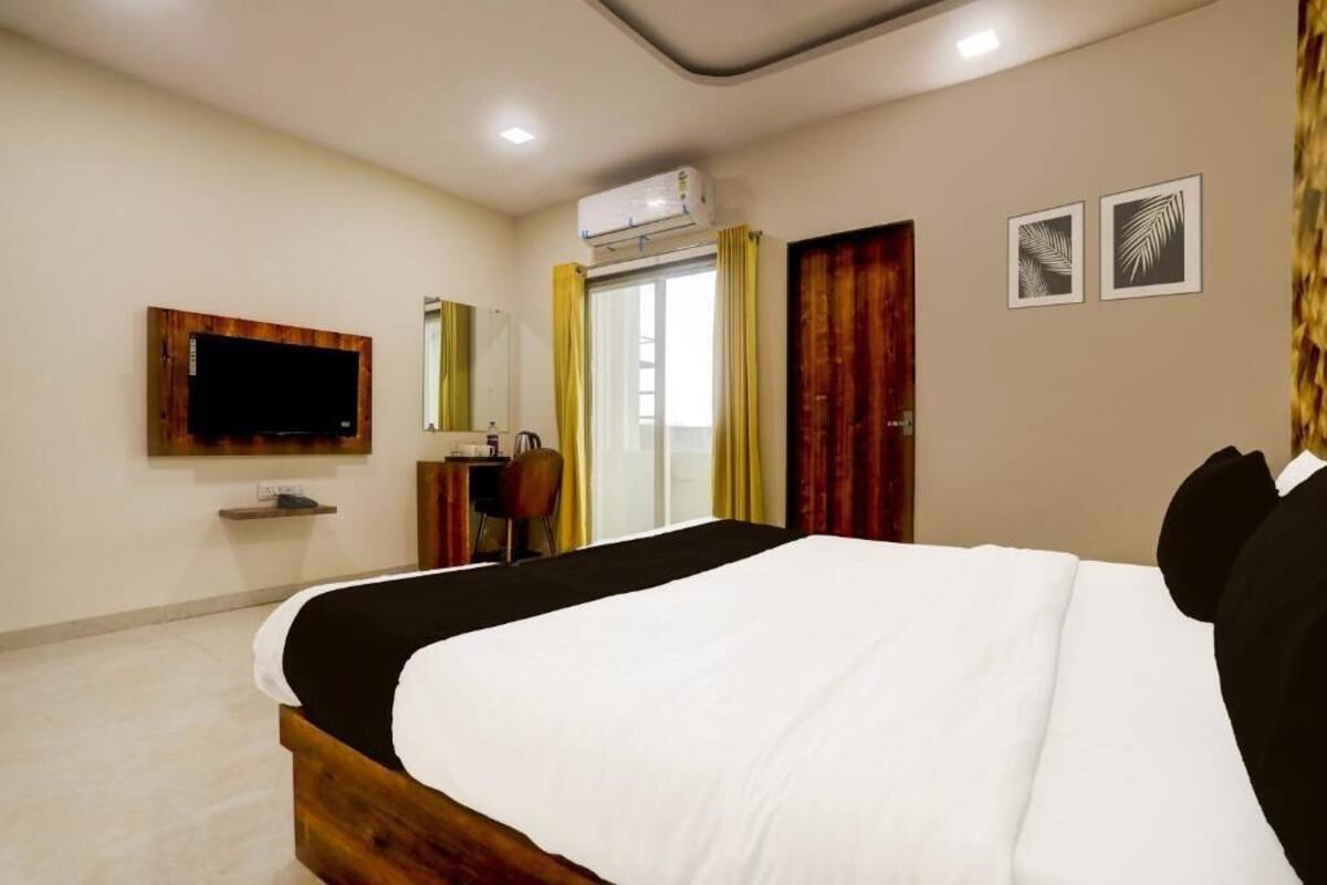 A hotel room with a large bed, a flat-screen TV mounted on the wall, an air conditioning unit, a desk with a chair, and two framed pictures on the wall.
