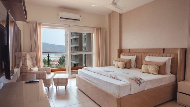 Habitación de hotel con una cama grande, televisión, sofá y balcón con vistas a edificios y montañas.